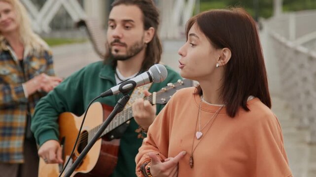 Medium shot of artistic teenage girl with red hair singing emotional song with expressive hand gestures, accompanied by male companion with guitar, while woman is enjoying and clapping in background
