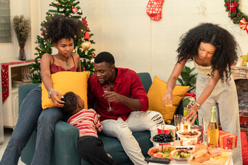 A Black family enjoys a cozy Christmas or New Year's Eve party at home. The African American parents and their young child relax on a sofa. Togetherness and holiday celebration in the living room.