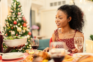 A beautiful young Black woman is sitting thoughtfully at a festive table during a Christmas dinner with family or friends at home.