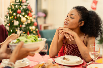 A beautiful young Black woman is enjoying a Christmas dinner with family or friends at home. She sits thoughtfully at a festive table decorated with food and wine