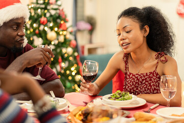 Black couple enjoys a festive Christmas dinner with friends or family. The African American woman holds a glass of red wine, talking and smiling. Holiday party with food, celebration