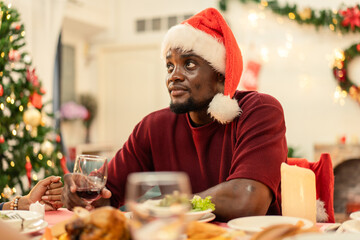 Thoughtful Black man wearing a Santa hat holding wine at a festive Christmas dinner. He is seated at the holiday table with food. Seasonal moment of reflection and celebration at home