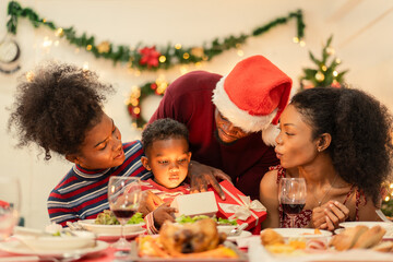Joyful Black family gathered at the Christmas dinner table watching a boy open a glowing red gift box. Magic of Christmas, festive excitement, and happy family bonding during the holiday season