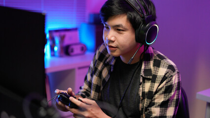 Attractive Asian male gamer enjoys playing online games on his computer feeling happy while live...