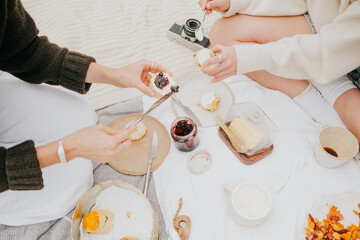 Two women are sitting on a blanket and eating food. One of them is holding a camera. The food on the table includes a cake, a pie, and a sandwich
