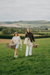 Naklejka premium Two women are strolling through a lush green field, each carrying a basket and a bottle. The scene is calm and tranquil, with the women enjoying the beauty of nature around them