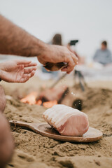A man is seasoning a piece of meat on a wooden board on a beach. The scene is casual and relaxed, with the man enjoying the outdoors while preparing his meal