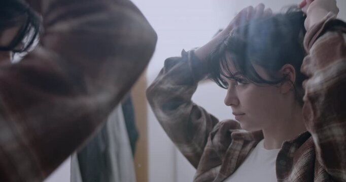 Young woman puts her hair up in a ponytail in a mirror in her bedroom and puts in her wireless headphones. Getting ready to go out with friend.