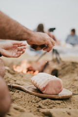 A man is seasoning a piece of meat on a wooden board on a beach. The scene is casual and relaxed, with the man and others around him enjoying the outdoors