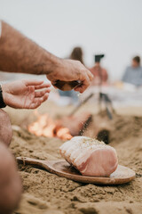 A man is seasoning a piece of meat on a wooden board on a beach. The scene is casual and relaxed, with a group of people nearby