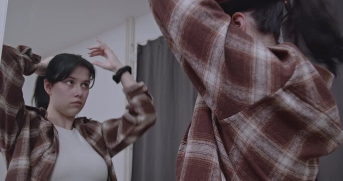 A young woman ties her hair up while looking in the mirror, getting ready to start her day at home.