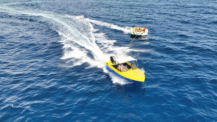 Aerial drone tracking photo of powerboat towing inflatable tubes used for water sports activities in deep blue open ocean island sea