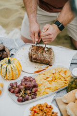 A man is cutting a large piece of meat on a table with a variety of food items