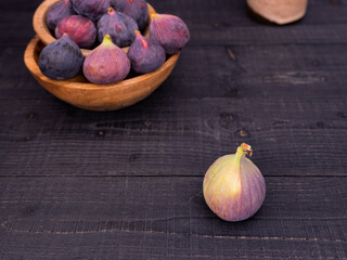 Fresh figs displayed on a black wooden table with one in front and more in wooden bowls, rustic and moody composition with vase in background