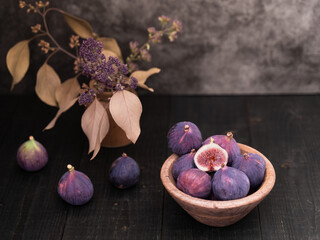 Half sliced fig in focus with whole figs nearby in a wooden bowl on dark table, rustic still life with vase decor in background