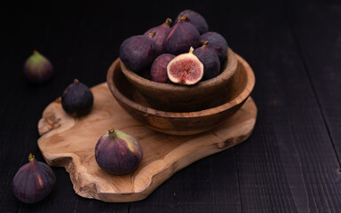 Half sliced fig in stacked bowls on wooden board with whole figs placed diagonally toward background, moody dark still life