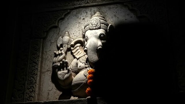 Intricate stone carving of Ganesh adorned with marigold flowers in shadow
