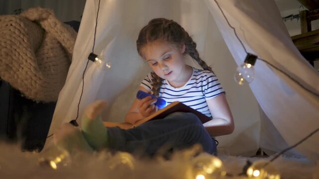 girl reading in cozy tent by flashlight and string light inside bedroom while child holds book and smiles during indoor bedtime reading with soft cozy light and warm tent atmosphere and gentle glow