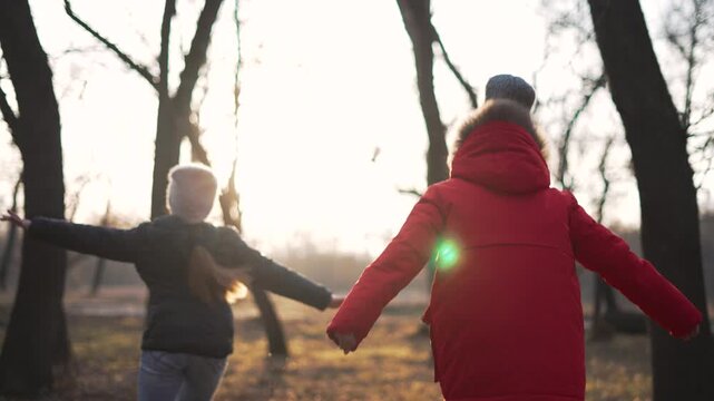 Two kids run in park among bare tree and leaf litter at winter dusk with sun flare lighting knit hat and red coat as girl and child play and walk along forest path in gentle motion and laughter