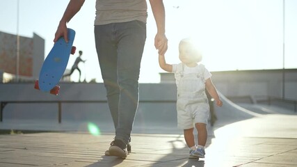Father holding toddler hand and walking through skatepark park area while carrying skateboard beside them parent and child in cap and overalls sun flare and urban ramp show warm playful family bonding