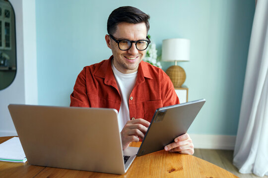 Young man engaged in digital work at home, using tablet and laptop while sitting at wooden table in modern living space - Powered by Adobe