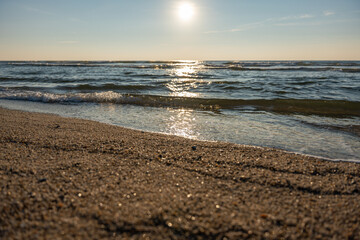 Golden sunlight sparkles on gentle sea waves along a sandy beach. The horizon glows softly under the evening sky.