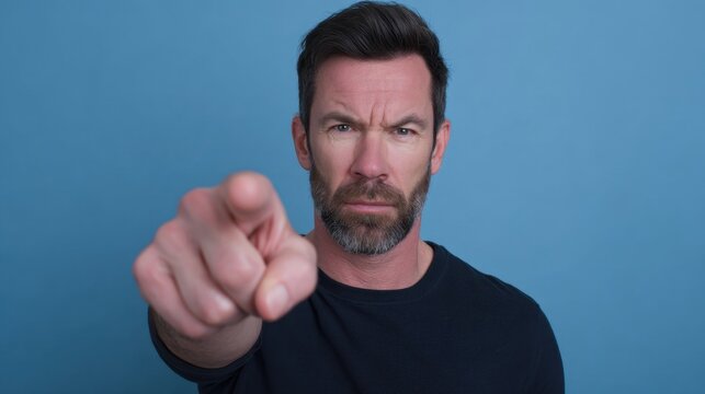 Serious man with beard pointing finger towards camera on blue background expressing authority and confidence in studio setting