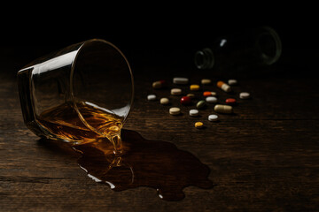 Glass of whiskey tipped over spilling alcohol on rustic wooden table scattered pills and empty bottle in background dark dramatic scene addiction concept