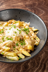 Golden French fries topped with creamy sauce, shredded cheese, and fresh herbs in a rustic bowl on wooden surface. Savory snack with rich texture and appetizing presentation.