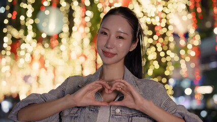 Portrait of happy Asian woman shows heart with fingers on evening street. Pretty Japanese lady expresses romantic feelings by gestures in night town decorated with light garlands