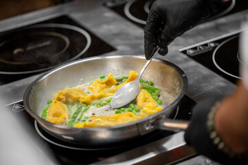 Chef in black gloves cooking stuffed pasta with peas in stainless steel pan on electric stove in professional kitchen. Close-up of food preparation and culinary technique.