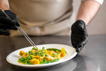 Chef plating gourmet pasta with green peas and asparagus, using tweezers for garnish. Professional kitchen setting, focus on culinary precision and fine dining presentation