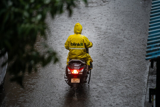 Blinkit delivery rider wearing a yellow raincoat drives a scooter on a wet road during heavy monsoon rain in Bangaluru 