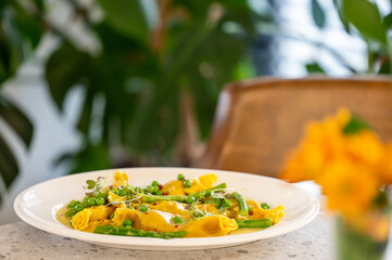 Gourmet stuffed pasta with yellow sauce, peas, asparagus, and microgreens served on a white plate in a cozy restaurant setting with floral decor and natural light.