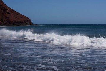 Fototapeta premium Dynamic ocean wave with foam and spray on the volcanic beach of Tenerife. Beautiful natural seascape showing strength, motion, and contrast of deep blue Atlantic water.