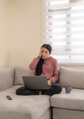 Naklejka premium A woman sitting cross-legged on a couch looks upset while viewing something on her laptop. Depicts emotional reaction, sadness, or unexpected news.