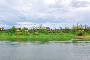 Calm river flows beneath a cloudy sky, reflecting soft light as it meets lush, green embankment dotted with trees and hidden homes. Tranquil scene evokes quiet charm and natural rustic harmony