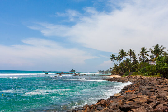 View of turquoise waves crashing against the rocky shore, palm trees swaying gently under a blue sky dotted with clouds, Coconut beach, Weligama, Sri Lanka.