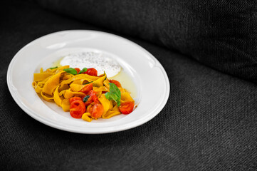 Tagliatelle pasta with cherry tomatoes, basil, and ricotta on a white plate over dark fabric. Rustic homemade meal with fresh ingredients and soft natural lighting.