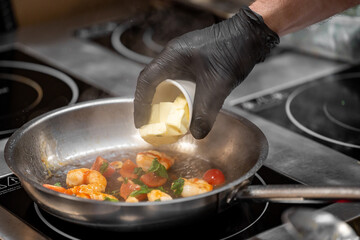 Chef adds butter to shrimp, cherry tomatoes, and herbs in a hot pan on electric stove. Close-up of cooking process in professional kitchen with steam rising.