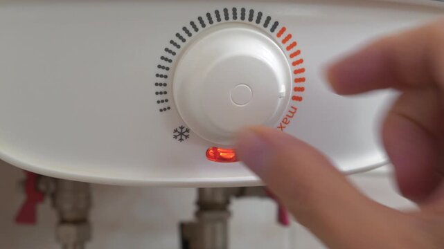 Regulator for heating cold water in boiler. Man turns temperature control on heater. Red light indicator comes on, boiler has turned on to heat water. Boiler heats water from cold water supply system