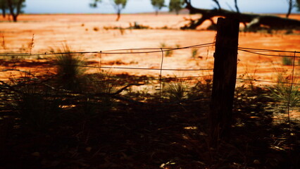 A sturdy wooden fence post supports tangled barbed wire against a vivid orange earth. Sparse vegetation dot the terrain, and distant green trees frame the expansive sky as sunlight filters down.