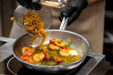 Chef pours cooked tagliatelle into pan with shrimp, cherry tomatoes, herbs, and garlic butter sauce. Close-up of seafood pasta preparation in professional kitchen.