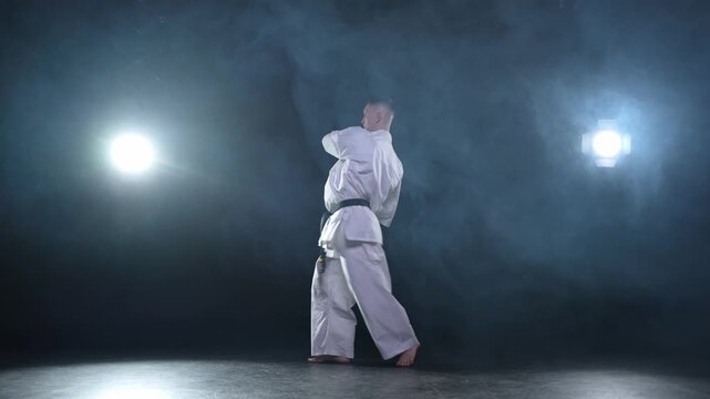 Strong man fighter in white kimono for karate martial arts with black belt preparing for fight training in black background. Man training moves punches kicks practicing shadow fight ready for sparring