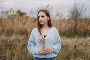 Candid outdoor portrait of a young woman standing in tall grass, holding dried reeds and gazing thoughtfully, natural light highlights authenticity and quiet credibility in the scene.
