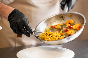 Chef plating shrimp tagliatelle pasta with cherry tomatoes and herbs in a professional kitchen. Culinary presentation, gourmet food, cooking process, restaurant concept.