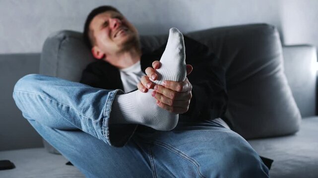 A young man has a foot injury.
A man hit his foot on a sharp object and is screaming in pain.
The man is holding his injured heel with his hand.