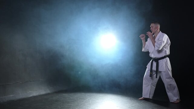 Strong man fighter in white kimono for karate martial arts with black belt preparing for fight training in black background. Man training moves punches kicks practicing shadow fight ready for sparring