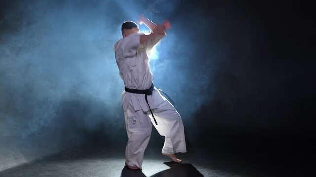 Strong man fighter in white kimono for karate martial arts with black belt preparing for fight training in black background. Man training moves punches kicks practicing shadow fight ready for sparring