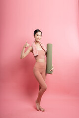 An Asian woman in sporty outfit with a yoga mat gives a thumbs up isolated on pink